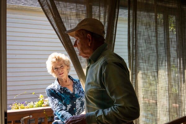 In a navy-and-white print blouse, Margaret Newcomb holds back a curtain while watching her husband Tim, in a ball cap and shirt.