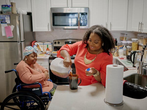 In a kitchen where a stainless steel refrigerator and microwave serve as a backdrop, Feylyn Lewis pours tea from a gallon jug into a brown mug while her mother, sits in a wheelchair behind her.