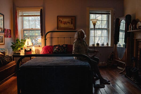 Dottye Burt sits on the edge of a bed in a darkened room, with a bedside table lamp behind her.